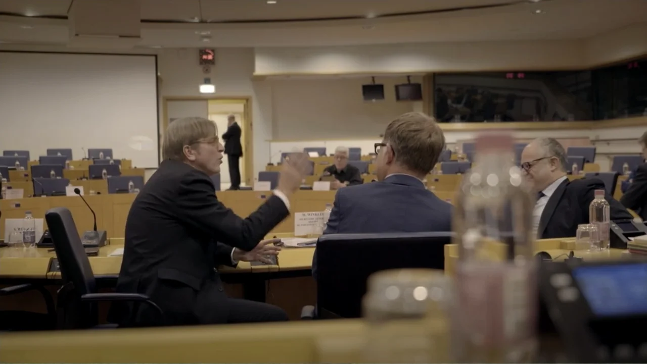 Brexit: Behind Closed Doors (2019) movie still: medium — A formal legislative chamber features rows of blue seats and light wood desks. Two men sit…