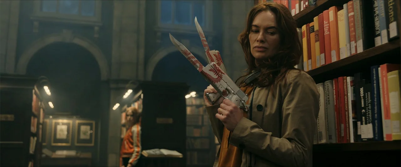 Gunpowder Milkshake (2021) movie still: medium — A dimly lit library features tall wooden shelves packed with books and stone arches. A woman stands…