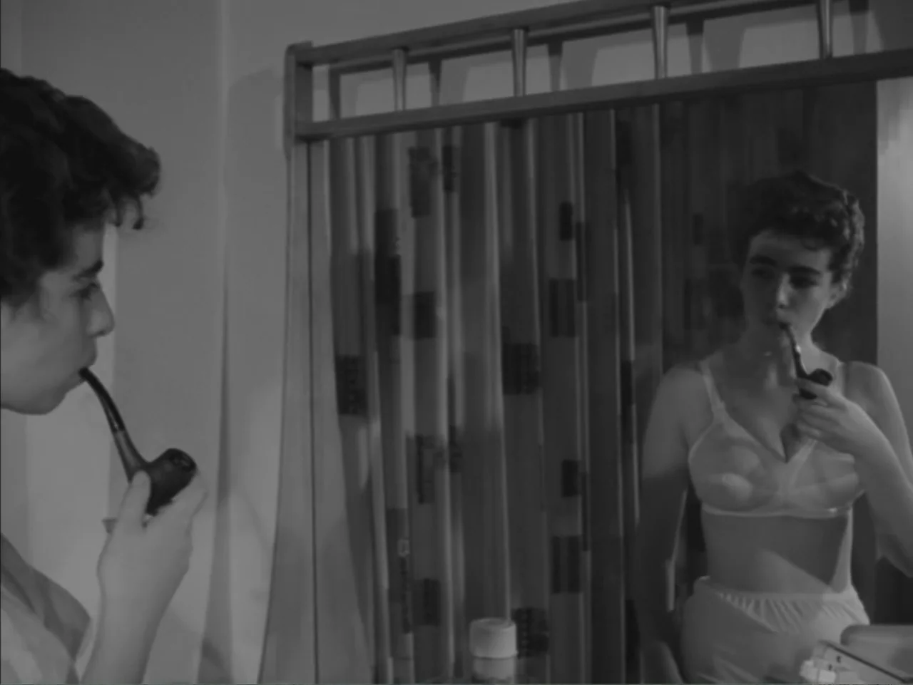 Chained Girls (1965) movie still: medium — A 1950s style domestic interior featuring a mirror and patterned curtain. A woman in undergarments…