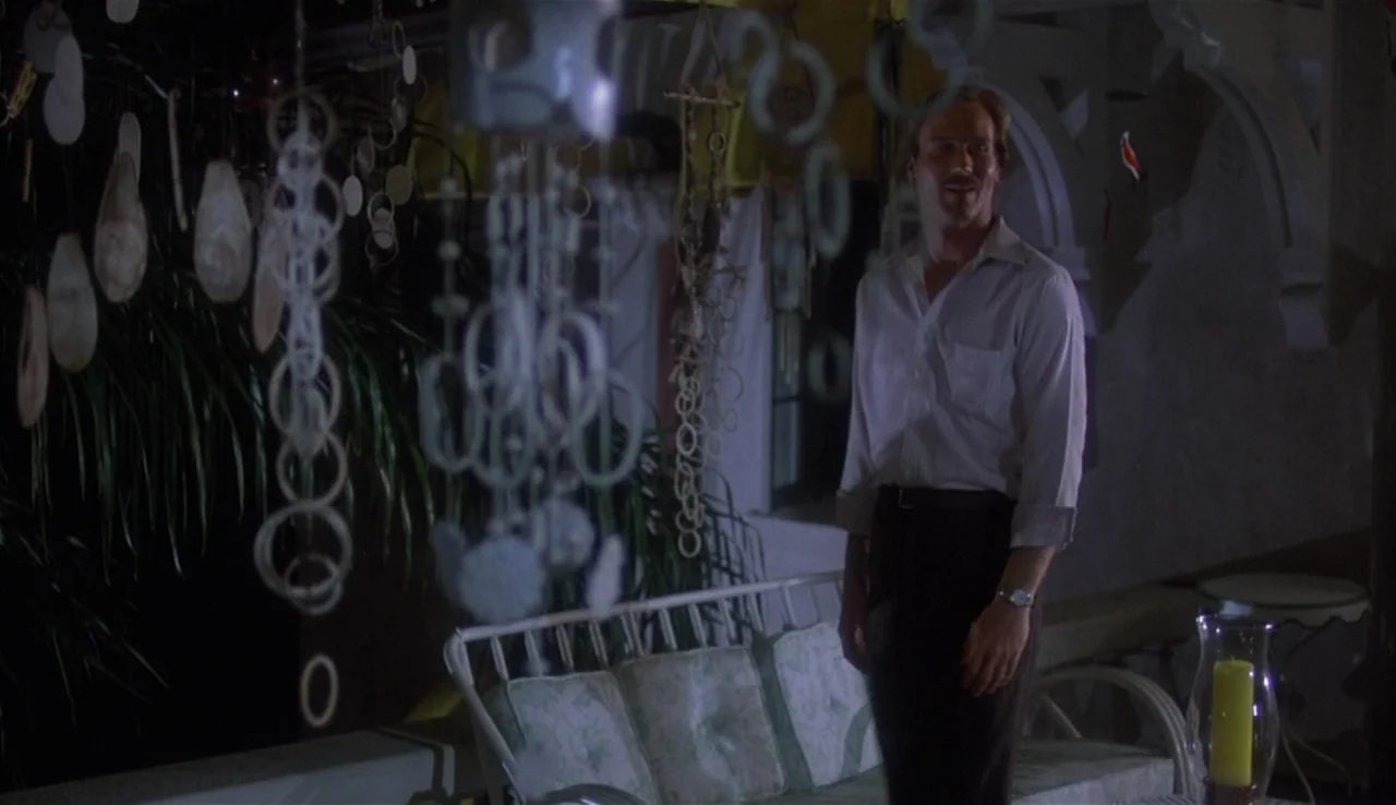 Body Heat (1981) movie still: medium — A dark outdoor patio at night featuring hanging decorative shell chimes in the foreground. A man stands on…