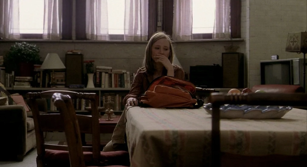 Birth (2004) movie still: medium — A quiet dining area features a table with a patterned cloth and an orange backpack. A young woman sits at the…