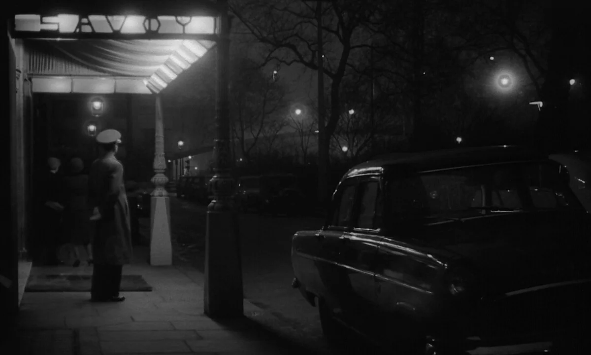 Night Of The Demon (1957) movie still: wide — A dark night scene outside a building entrance with a lit Savoy marquee sign. A lone doorman in a…