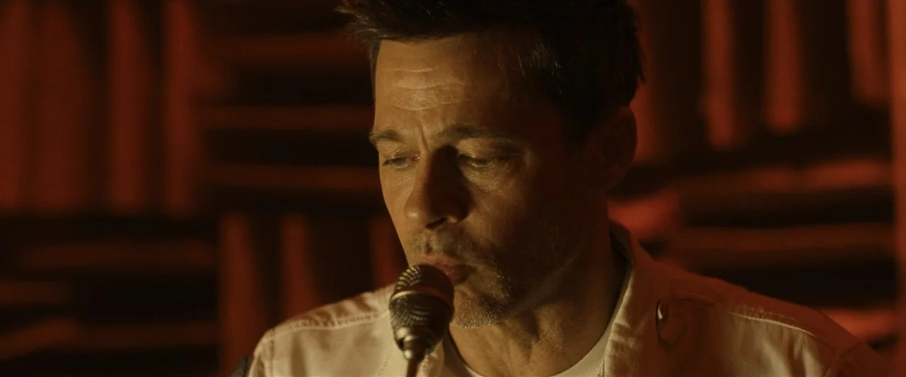 Ad Astra (2019) movie still: closeup — A dark recording studio features soundproofing panels on the walls in soft orange light. A man wearing a…