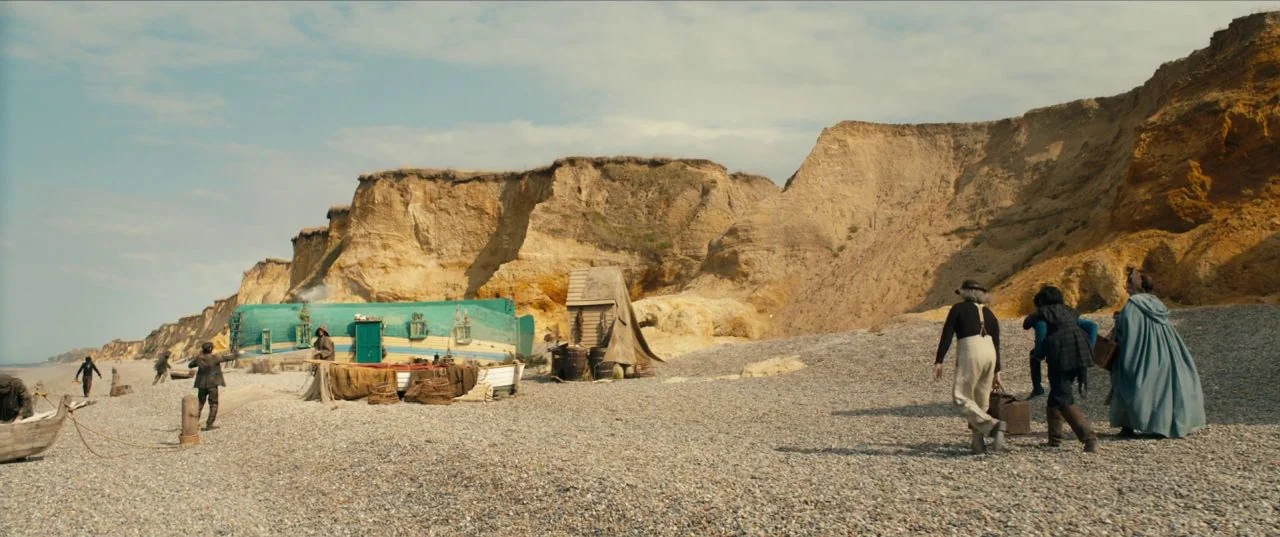 The Personal History of David Copperfield (2019) movie still: wide — A pebble beach stretches beneath high yellow cliffs under a bright sky. Several…