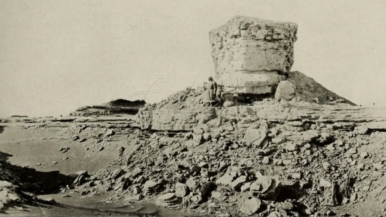 The Atlantis Puzzle (2024) movie still: wide — An arid, rocky landscape features a prominent natural stone pillar rising from the ground. A single…