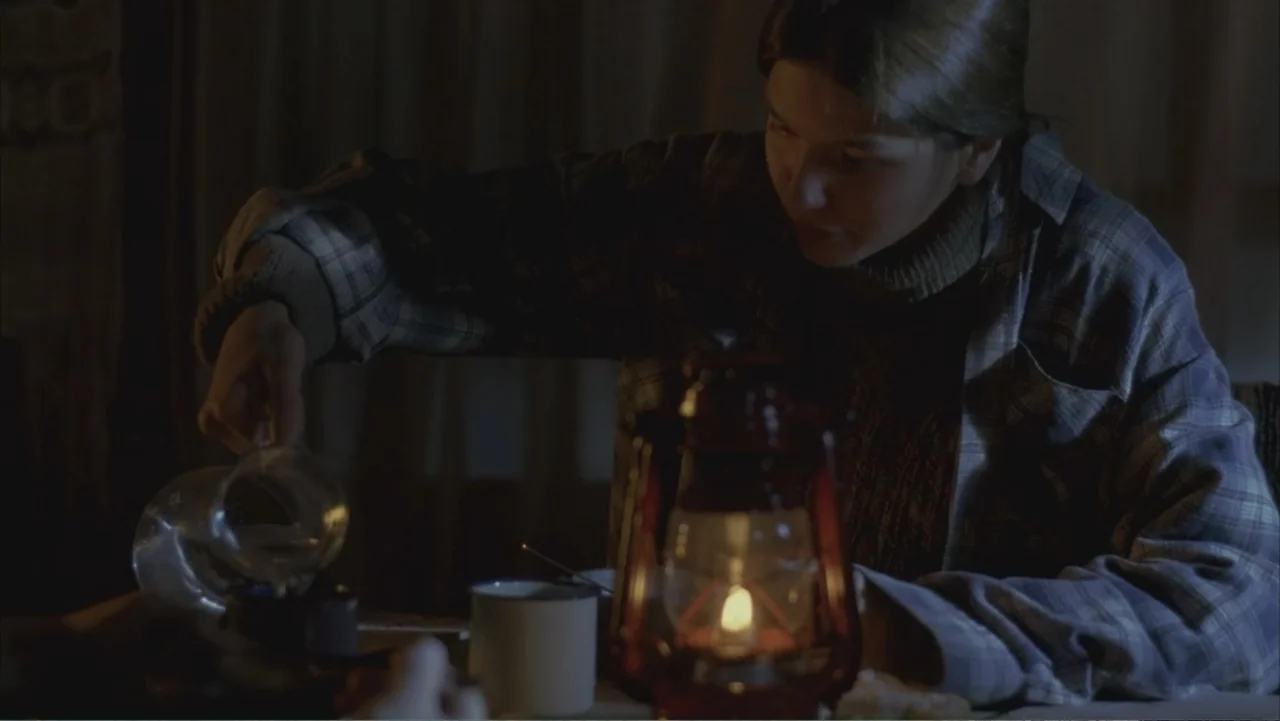 A Distant Place (2020) movie still: medium — A dimly lit room features a wooden table at night with simple rustic furnishings. A young woman in a…