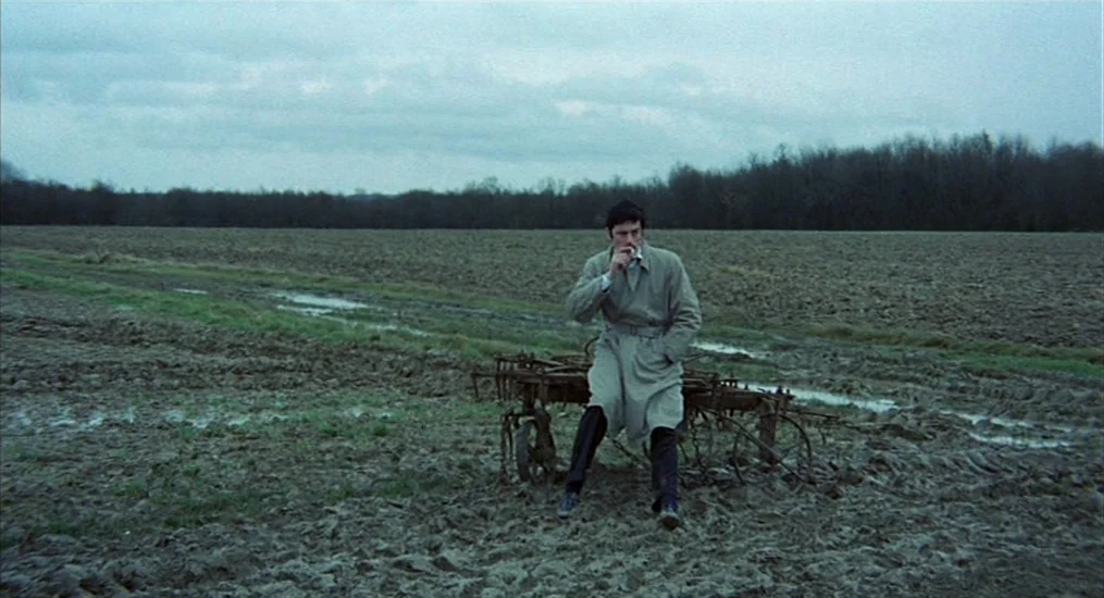 Le Cercle Rouge (1970) movie still: wide — A vast, damp, ploughed field sits under an overcast sky with a forest line. A man in a trench coat sits…