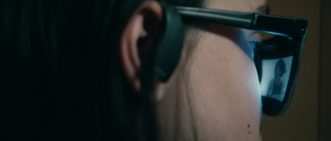 The Purge (2013) movie still: closeup — A dark environment holds a person wearing glasses with internal reflective displays. The person stares…