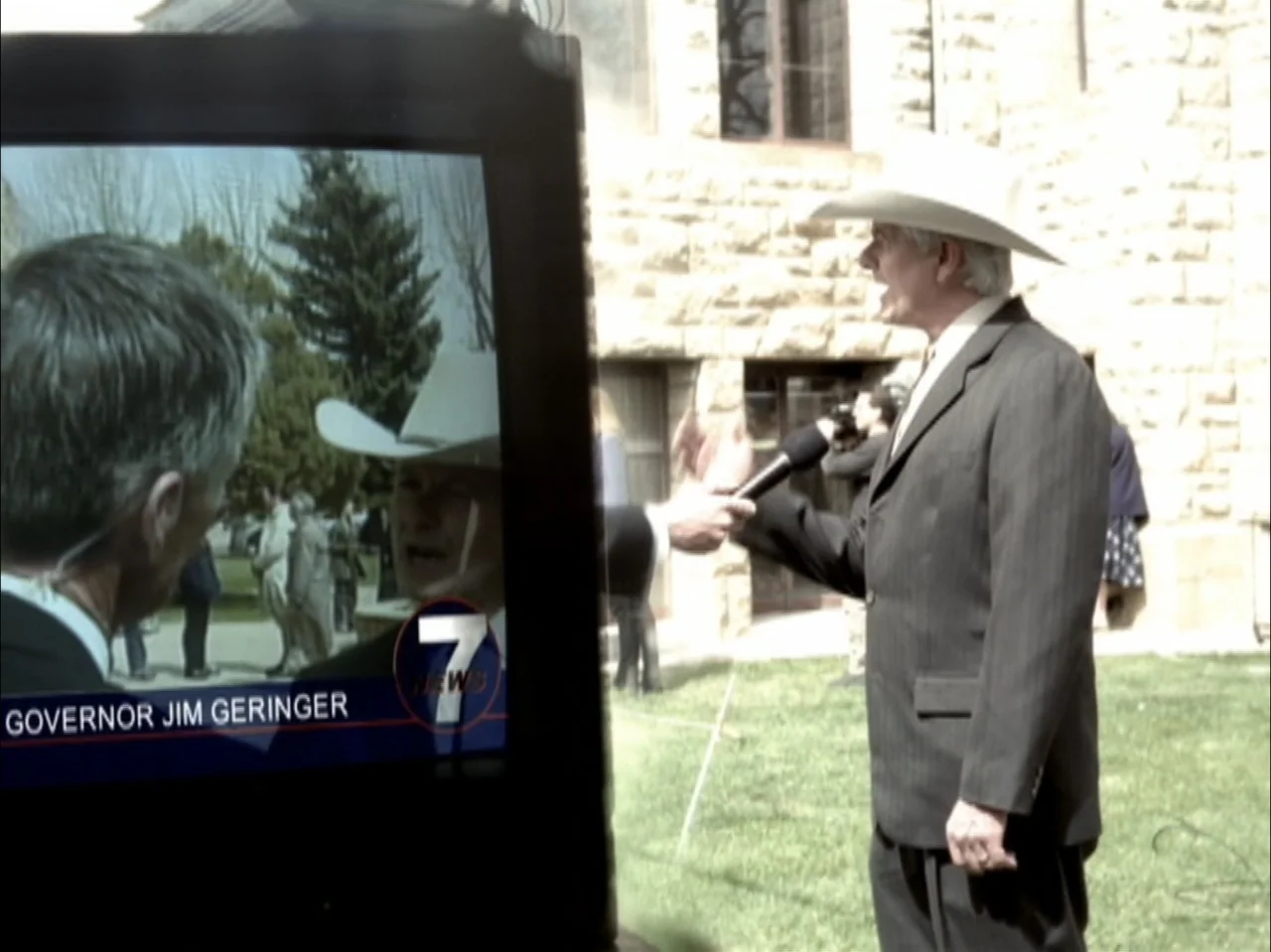 The Laramie Project (2020) movie still: medium — A man wearing a cowboy hat and suit speaks to a microphone outside a stone building. A television…