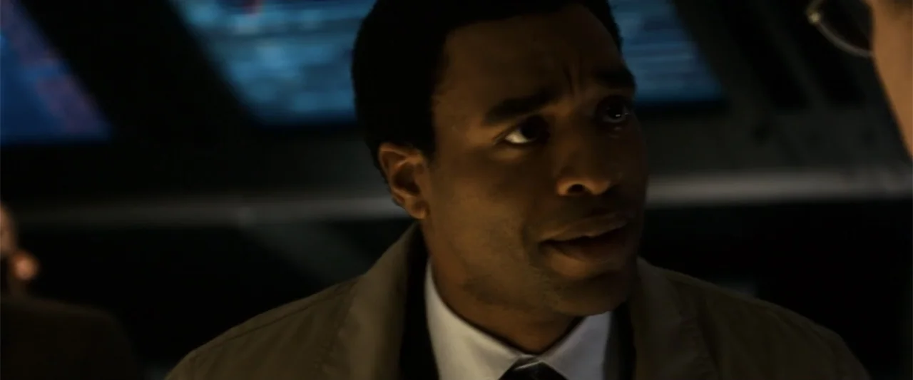 2012 (2009) movie still: closeup — A dimly lit interior space with glowing data displays in the background. A man in a trench coat is seen from the…