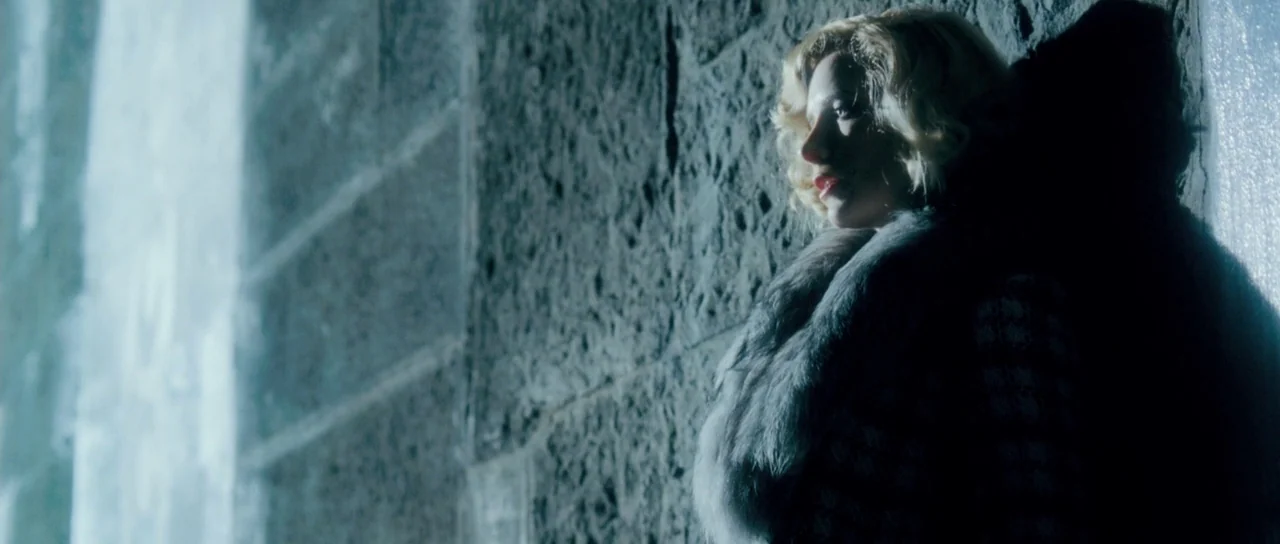Filth (2013) movie still: medium — A woman stands in the dark shadows next to a textured stone wall exterior. She leans against the surface while…