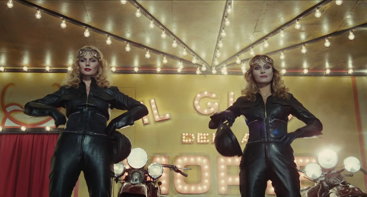 City of Women (1980) movie still: medium — Two women in black leather jumpsuits stand beneath a lit marquee at a carnival. They pose in front of…