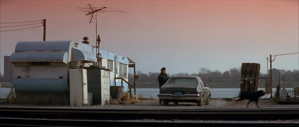 True Romance (1993) movie still: wide — A gravel bank borders a body of water near a faded mobile home structure. A person stands by an idling car…