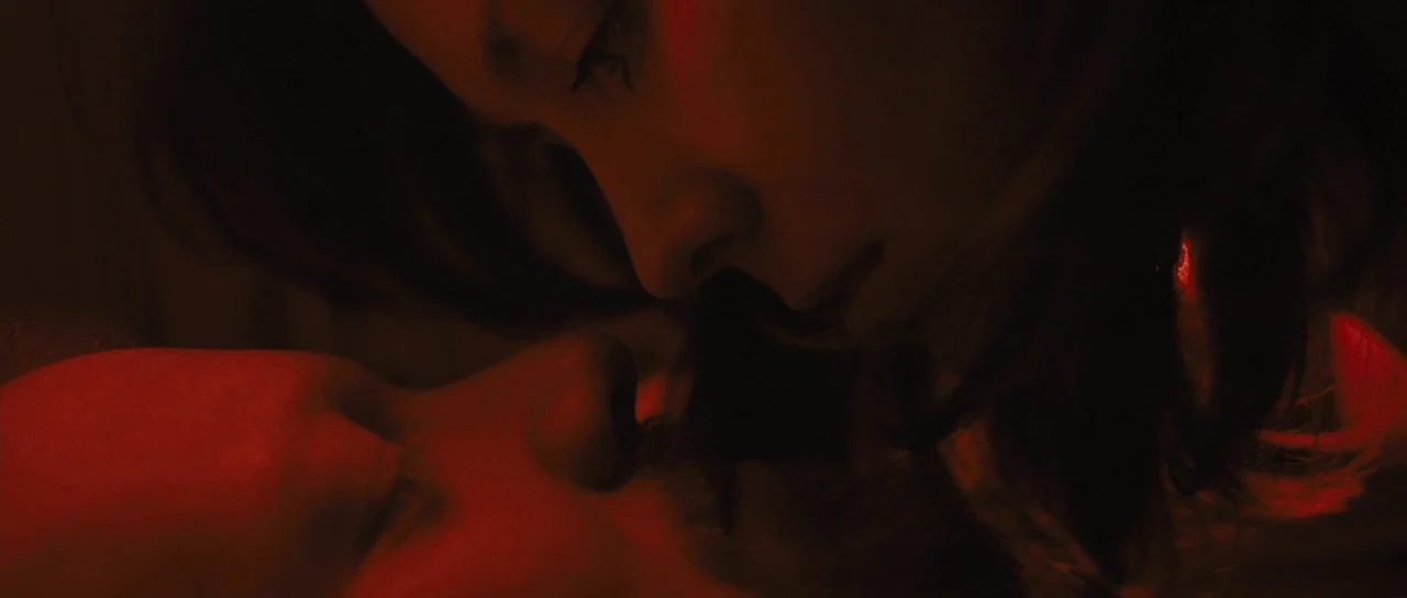 Sightseers (2012) movie still: closeup — A dark indoor space is illuminated by an intense red light source. A woman hovers over a man with a bearded…
