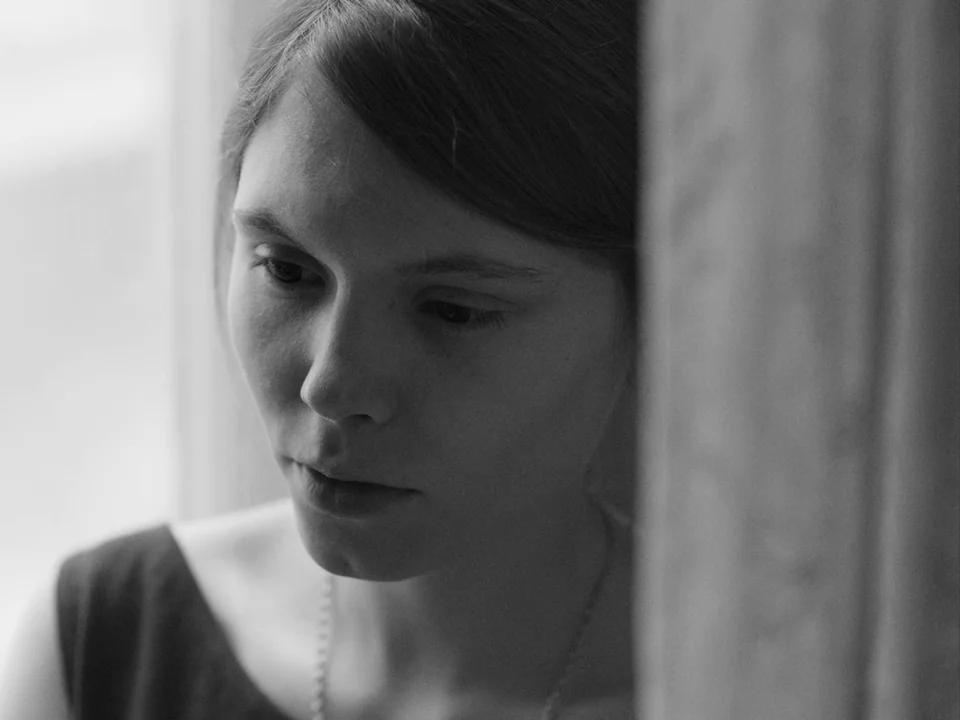 Ida (2013) movie still: closeup — An interior space features a young woman positioned near a window and wall. The woman gazes downward while leaning…