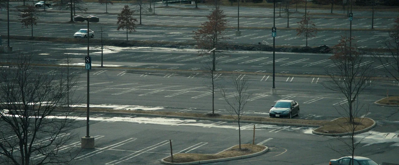 Compliance (2012) movie still: establishing — A vast, empty asphalt parking lot is marked by orderly white lines and light poles. Sparse leafless…