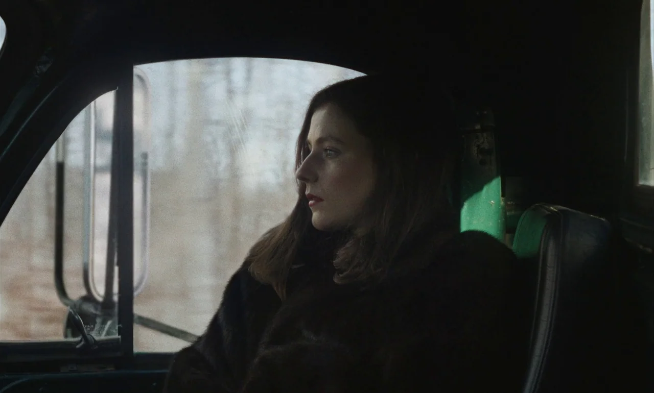 Eileen (2023) movie still: medium — The interior of a vehicle shows a woman sitting in the passenger seat. Outside the window, blurry trees suggest…