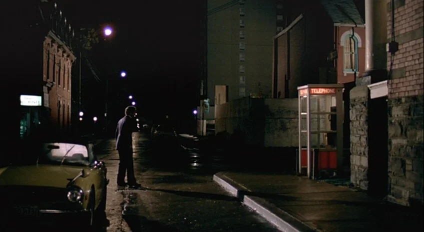 The Silent Partner (1978) movie still: wide — A dark city street at night features a yellow vintage car and a phone booth. A man in a dark suit…