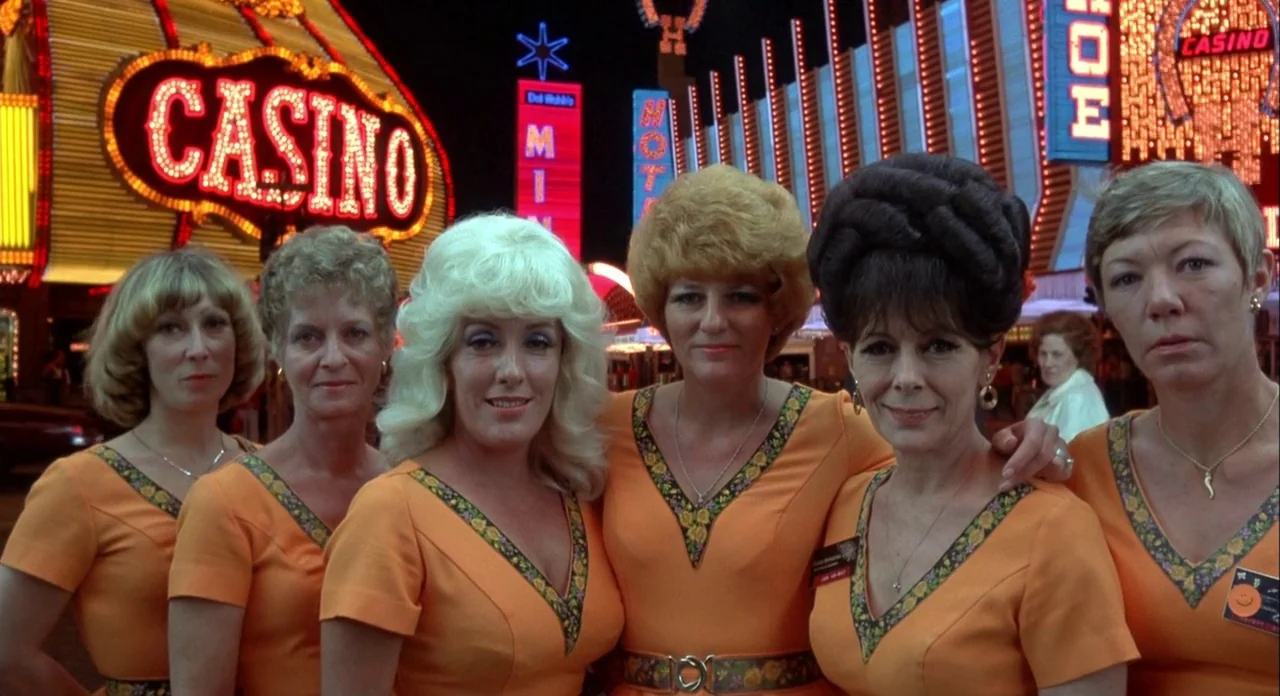 Koyaanasquatsi (1982) movie still: medium — Six women in matching orange uniforms stand in a row on a busy street. Brightly glowing neon signs for a…