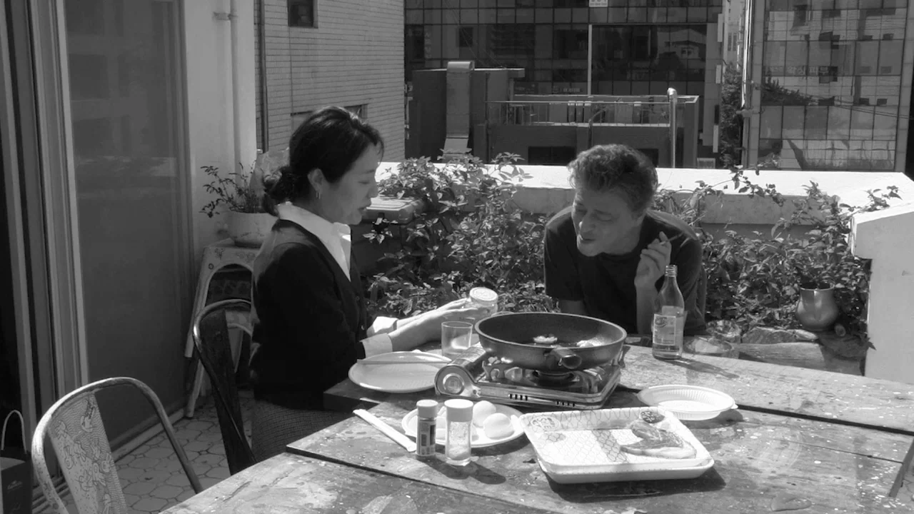 Walk Up (2022) movie still: medium — A rooftop terrace featuring a weathered wooden table with cooking equipment and greenery. A man and woman sit…