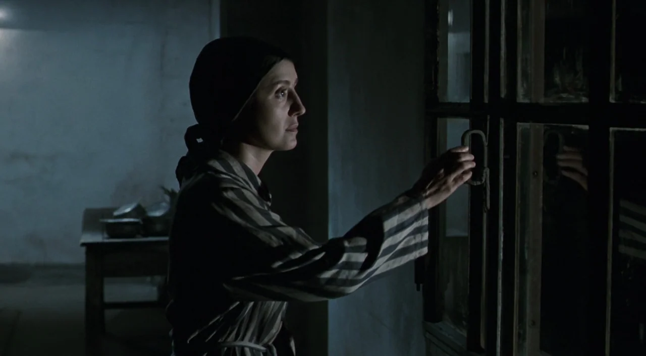 Life is Beautiful (1997) movie still: medium — A dimly lit room with bare walls and a wooden table in the back. A woman in a striped uniform stares…