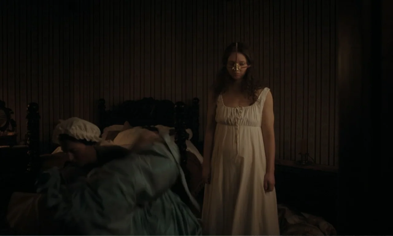 The Ugly Stepsister (2025) movie still: medium — A dark bedroom with vintage wallpaper features a woman standing by a carved wooden bed. A second…