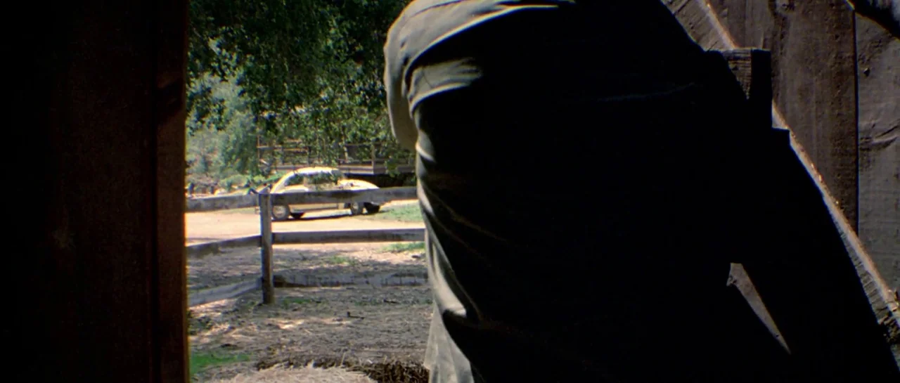 Friday the 13th Part III (1982) movie still: medium — A dark wooden barn interior frames a view of a bright rural landscape. A vintage sedan is…