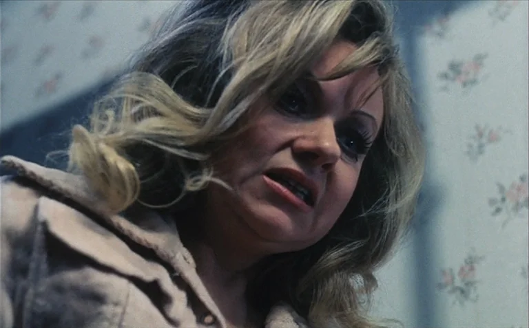 Business is Business (1971) movie still: closeup — A room with patterned floral wallpaper serves as the setting for this shot. A blonde woman in a…
