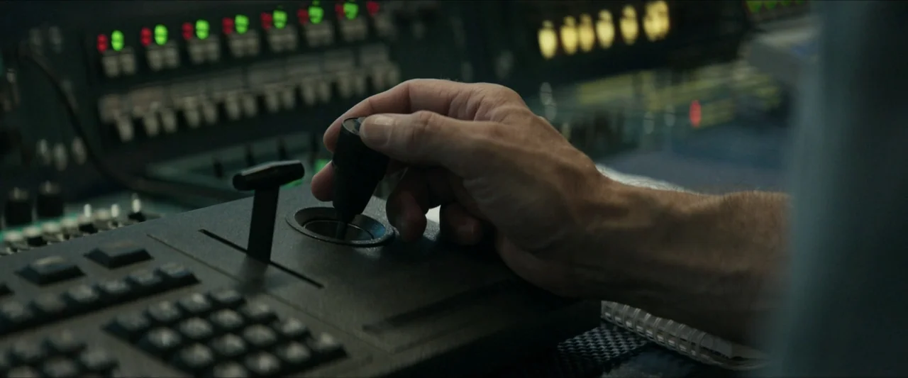 Last Breath (2025) movie still: closeup — A close view of a hand operating a technical joystick on a dark console. The background shows illuminated…
