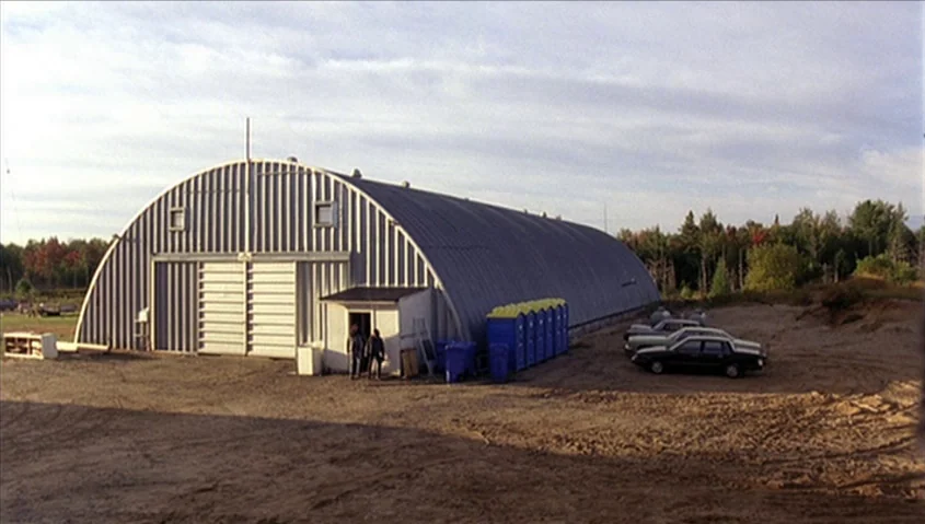 Dance Me Outside (1994) movie still: wide — A metal Quonset hut stands in a rural, sandy area with surrounding trees. Two men stand outside the…