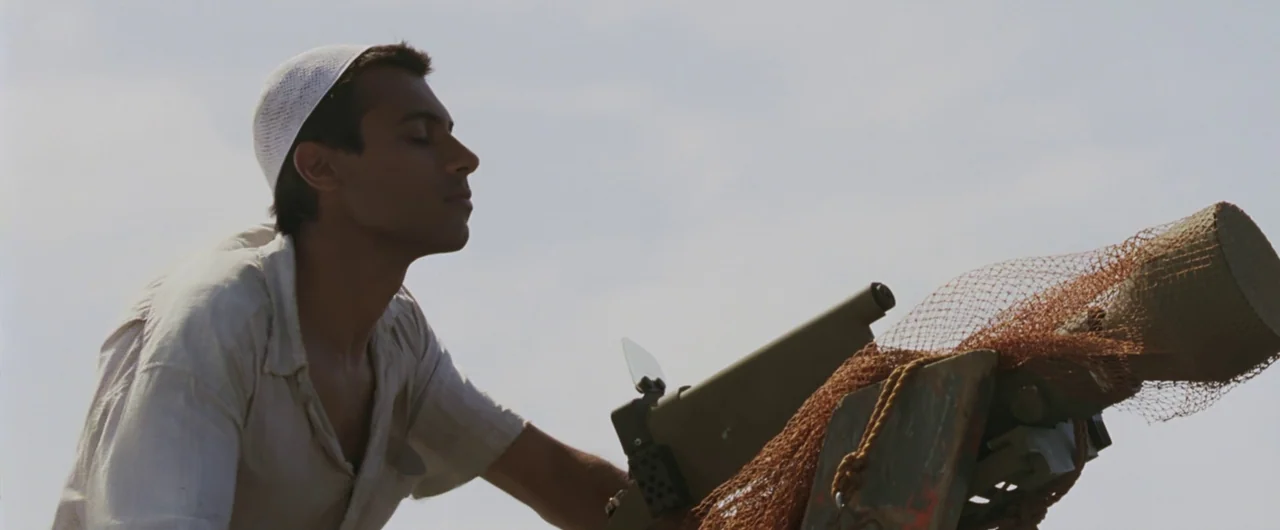 Syriana (2005) movie still: medium — An open field under a bright, overcast sky serves as a backdrop. A young man wearing a white skullcap adjusts a…