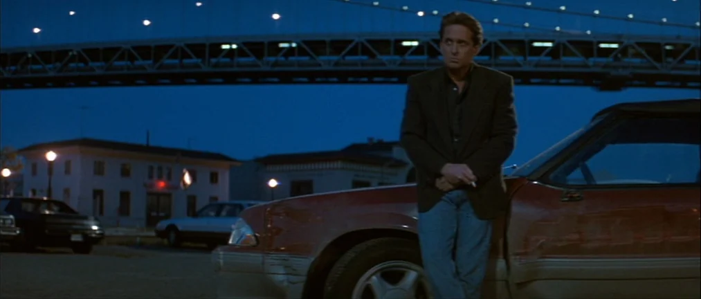 Basic Instinct (1992) movie still: medium — A dark parking lot sits beneath a massive suspension bridge during the blue hour. A man leans against a…