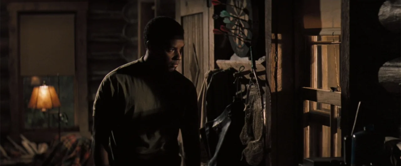 The Pelican Brief (1993) movie still: medium — A dimly lit log cabin interior features wooden walls and various hanging rustic equipment. A man in a…