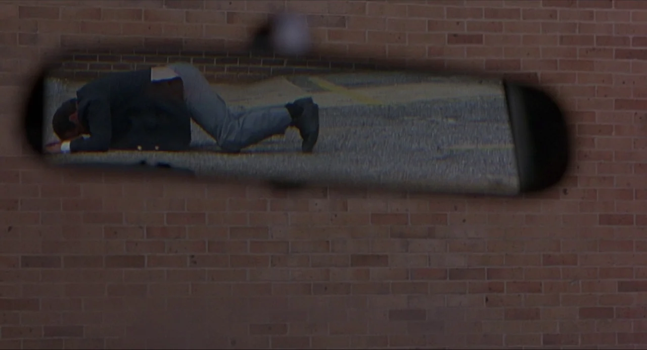 Serial Mom (1994) movie still: medium — A view through a rearview mirror shows a man kneeling on asphalt. A plain brown brick wall surrounds the…