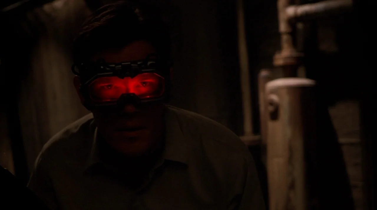 Hollow Man (2000) movie still: medium — A dark, cramped space filled with metallic pipes and industrial equipment. A man wearing red glowing goggles…