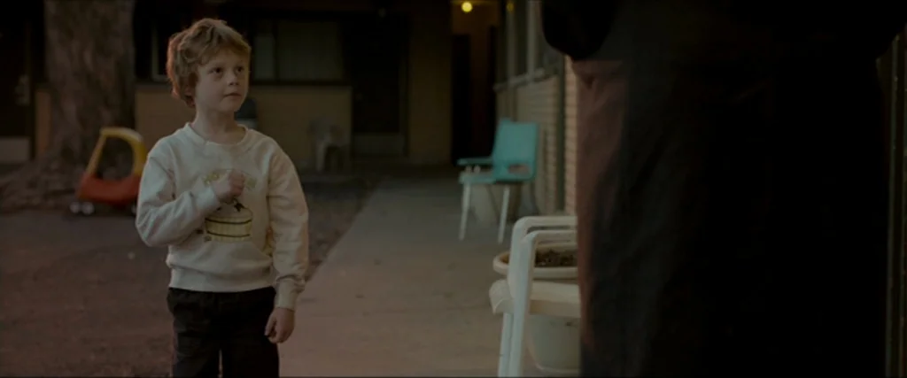 Clown (2014) movie still: medium — A dark outdoor walkway at an apartment complex features a concrete path and plastic chairs. A young boy faces an…