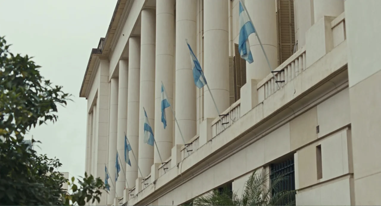 Belén (2025) movie still: establishing — A classical stone government building features tall columns and a balcony with flags. Multiple light blue…