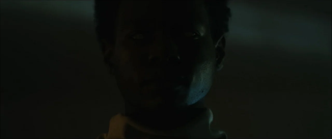 I'll Take Your Dead (2018) movie still: closeup — A darkened interior space with minimal light cast upon the subject. A man faces forward with his…