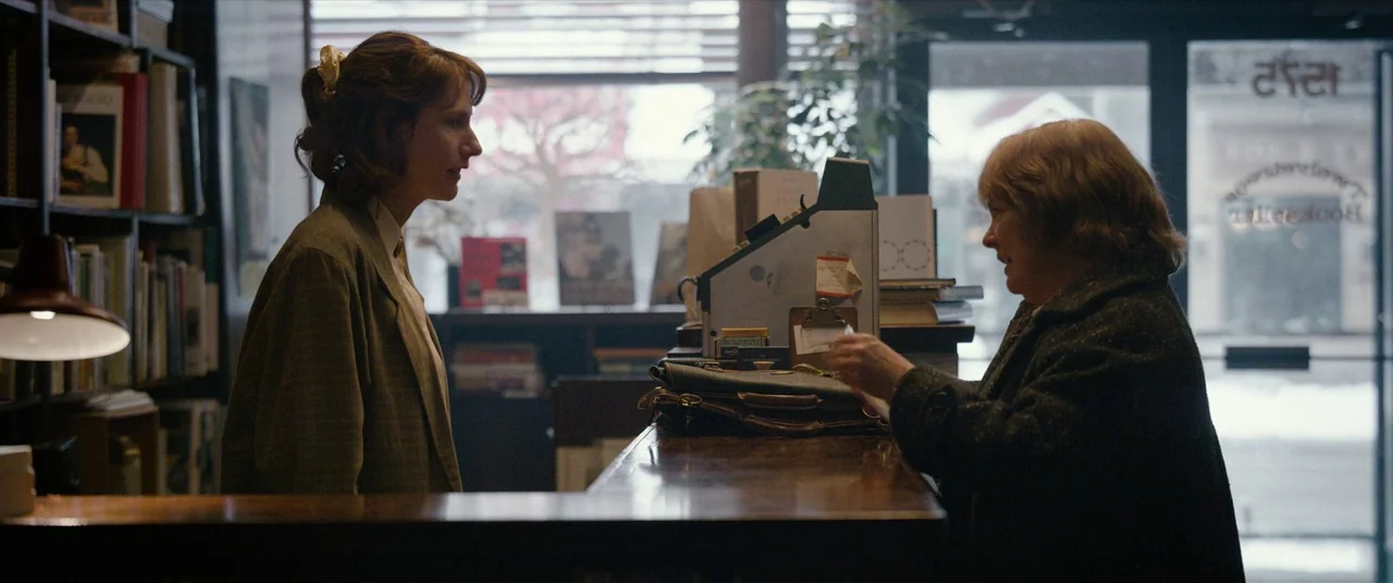 Can You Ever Forgive Me? (2018) movie still: medium — Two women stand on opposite sides of a wooden counter in a small shop. Bookshelves line the…