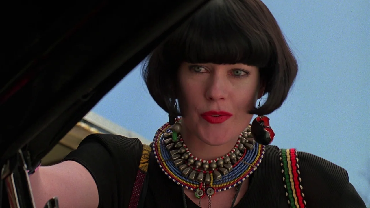 Something Wild (1986) movie still: medium — A woman stands in a bright outdoor space under a clear blue sky. She leans toward an open car door while…