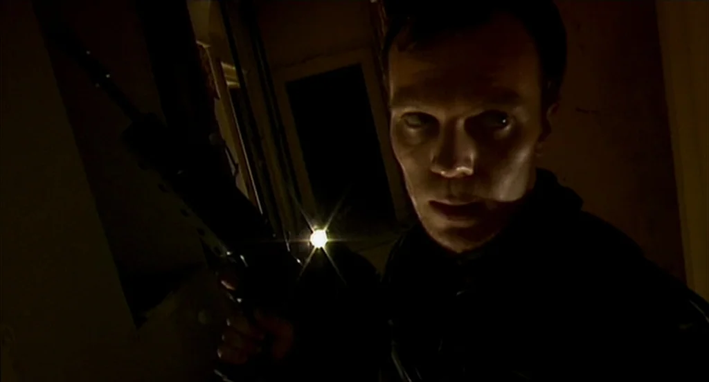 28 Days Later (2002) movie still: closeup — A dark, unlit interior space characterized by heavy shadow and indistinct architecture. A man holding a…