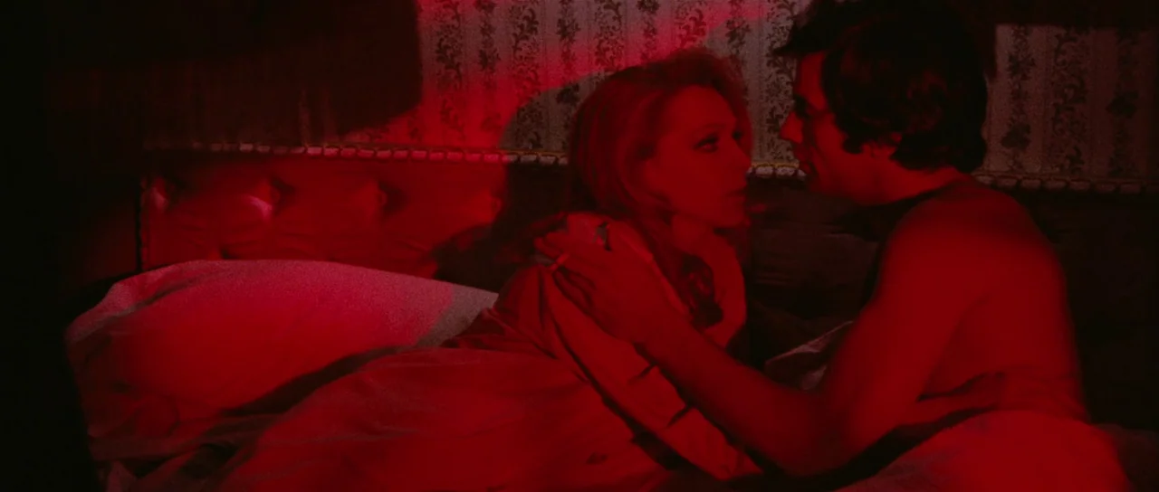 Death Walks On High Heels (1971) movie still: medium — A dimly lit room with patterned floral wallpaper and a cushioned headboard. A man and a woman…