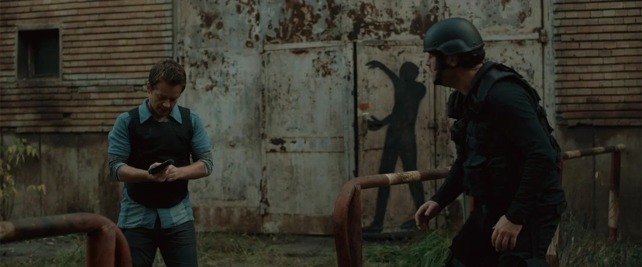 The Condemned 2 (2015) movie still: medium — An industrial area features a weathered brick wall and a faded metal door. Two men in tactical gear…