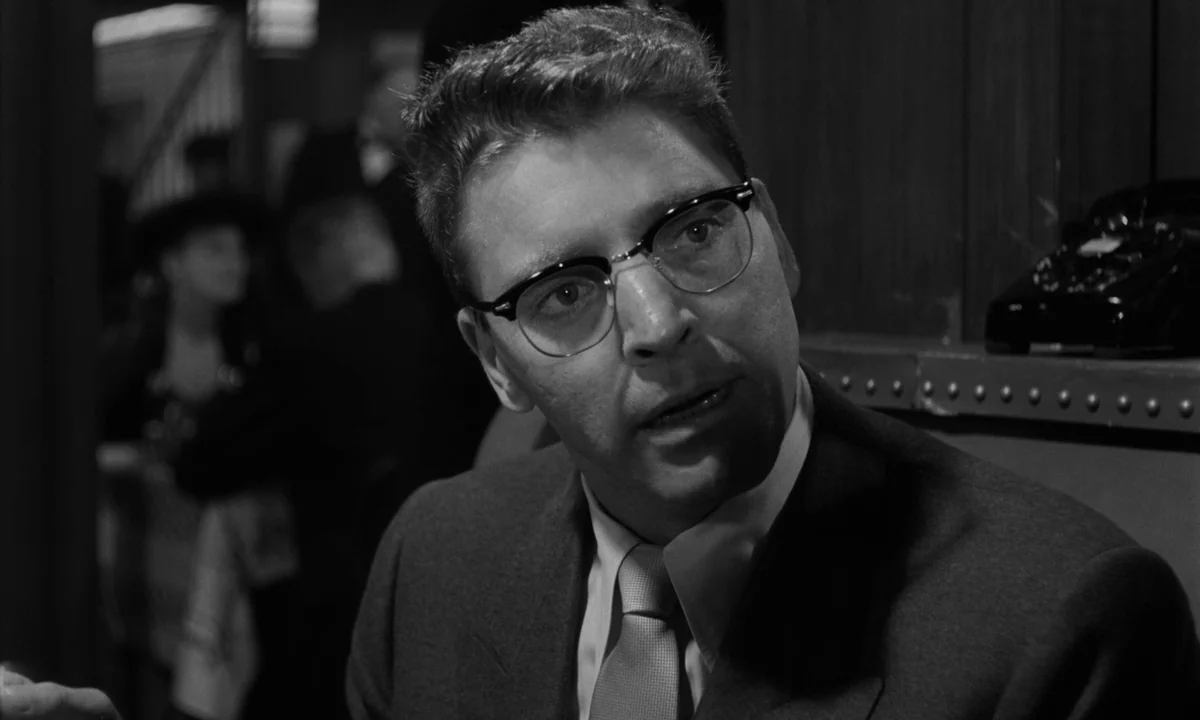 Sweet Smell of Success (1957) movie still: closeup — A dark, dimly lit room contains a man sitting at a desk. The man wearing eyeglasses looks…