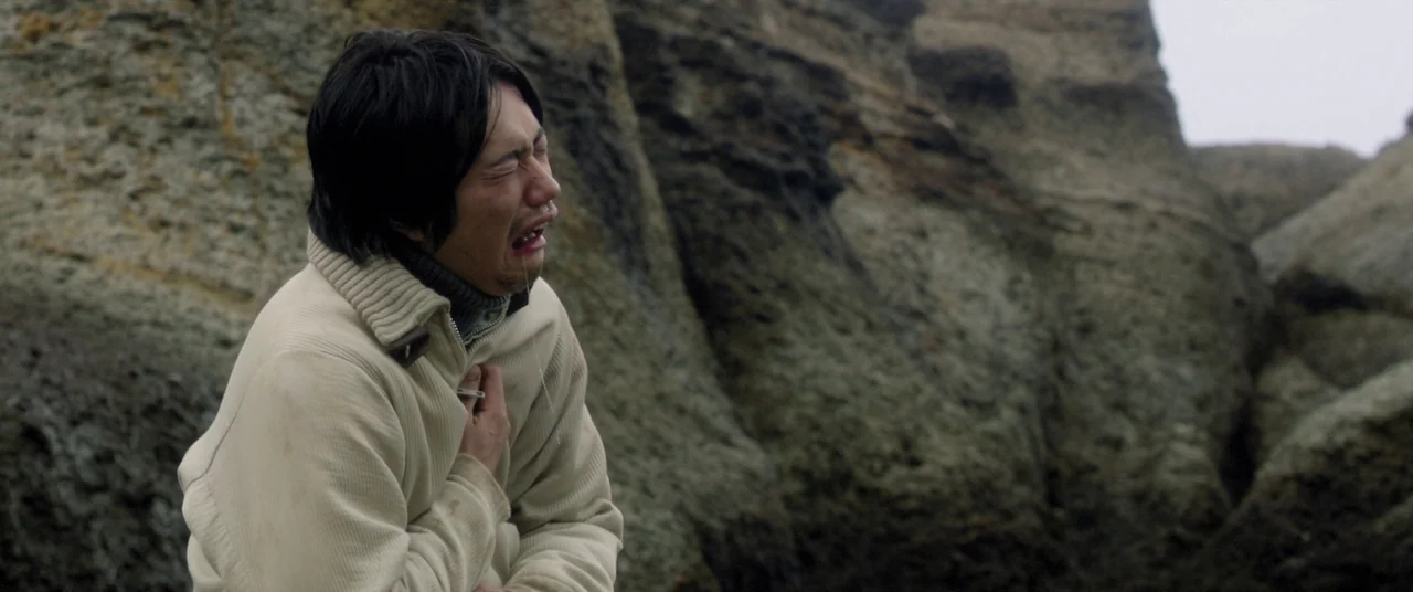 Norwegian Wood (2010) movie still: medium — A rocky coastal cliff with large layered stone formations under an overcast sky. A man in a…