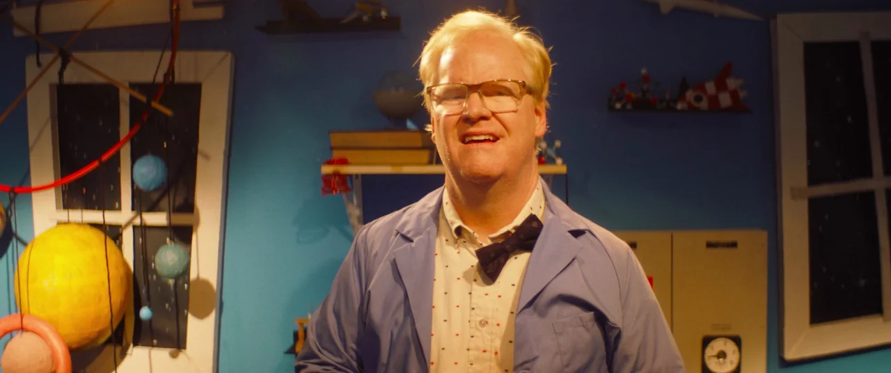 Linoleum (2022) movie still: medium — A bright blue room decorated with DIY solar system models and educational science props. A man in a lab coat…