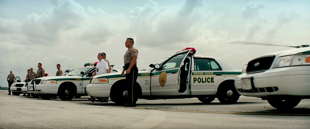 Pain & Gain (2013) movie still: wide — A row of white Miami-Dade police cruisers sits on a flat, gray paved tarmac. A group of uniformed officers…