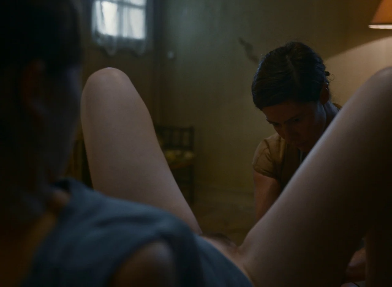 Happening (2021) movie still: medium — A dimly lit room contains two women in a private interaction near a bed. One woman wears a brown top while…