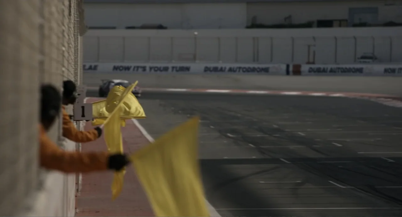 Gran Turismo (2023) movie still: medium — A concrete marshal post overlooks a winding asphalt circuit with blurred cars in distance. Two officials…