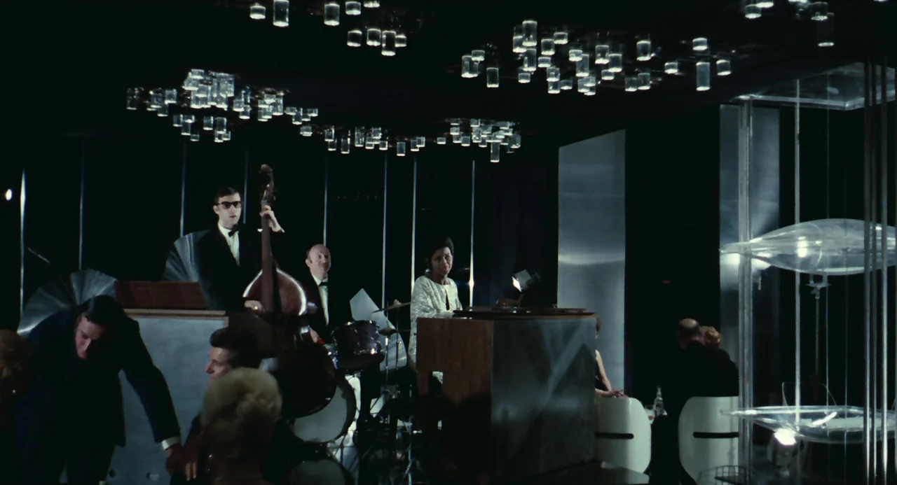 Le Samouraï (1967) movie still: wide — A dark, modern nightclub features a jazz band performing on a small raised platform. Multiple glass cylinder…